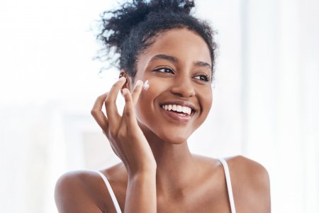 Smiling woman demonstrating how to apply cream to her cheek as part of her clear skin routine.